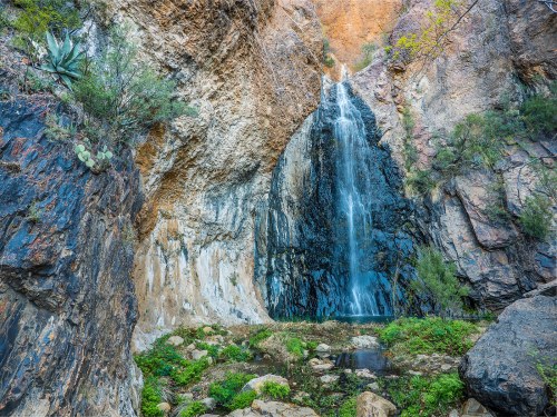 A serene waterfall cascades down rocky cliffs, surrounded by lush greenery and rugged terrain.