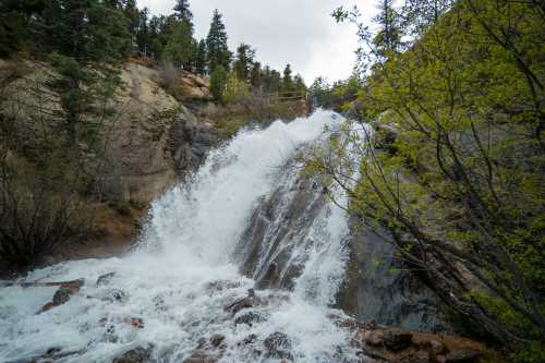 A cascading waterfall flows over rocky terrain, surrounded by trees and greenery under a cloudy sky.