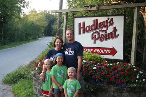A family of five stands in front of a sign for Hadley's Point Campground, surrounded by flowers and trees.