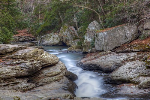 A serene stream flows between large rocks in a lush, wooded area, with autumn leaves scattered on the ground.