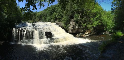 A scenic waterfall cascading over rocks, surrounded by lush green trees and a clear blue sky.