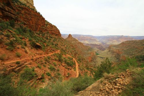 A scenic view of a canyon with red rock formations and a winding trail, surrounded by greenery and distant mountains.
