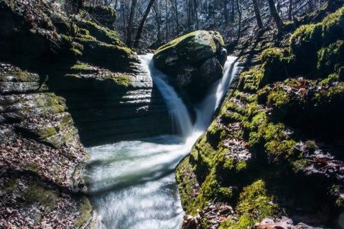 A serene waterfall cascades over moss-covered rocks in a lush, wooded area. Sunlight filters through the trees.