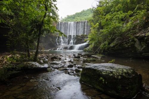 A serene waterfall cascades over rocks, surrounded by lush greenery and a calm river in a tranquil natural setting.