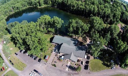 Aerial view of a building by a pond, surrounded by trees and parked cars on a sunny day.