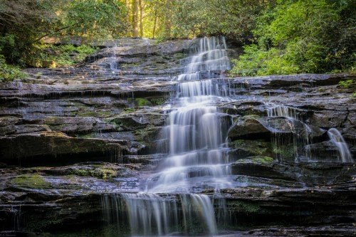A serene waterfall cascading over rocky ledges, surrounded by lush green foliage and dappled sunlight.