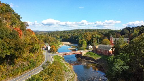A scenic view of a river winding through autumn-colored trees, with a bridge and buildings along the shore.
