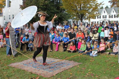 A performer walks a tightrope with a parasol, entertaining a large crowd of children and adults seated on the grass.