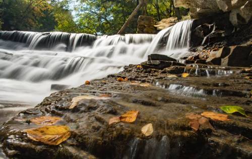 A serene waterfall cascades over rocks, surrounded by autumn leaves and lush greenery.