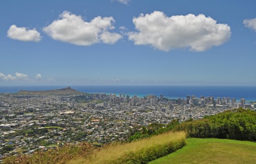 A panoramic view of a coastal city with skyscrapers, mountains, and a blue ocean under a partly cloudy sky.