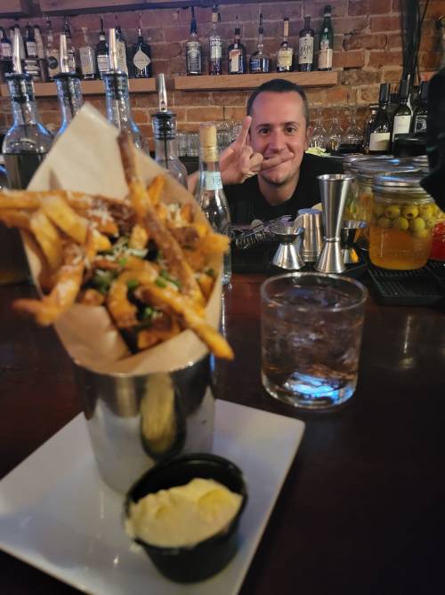 A plate of fries with dipping sauce in front of a bartender making a hand gesture, with drinks and bottles in the background.