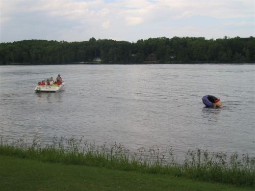A boat with people is on a lake, while someone in a life jacket leans over the water near the shore.