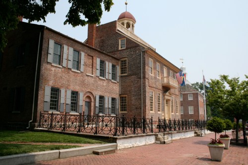 Historic brick building with a dome, surrounded by greenery and flags, featuring a wrought-iron fence and a brick walkway.