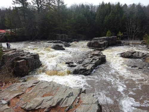 A rushing river flows over rocky outcrops, surrounded by trees and a cloudy sky. Two people stand on the shore.