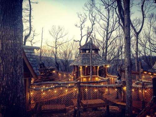 A whimsical treehouse surrounded by bare trees, adorned with string lights, at sunset.