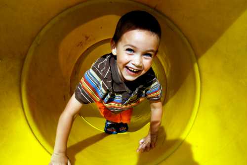 A smiling boy in a striped shirt climbs through a bright yellow slide tunnel, enjoying playtime.