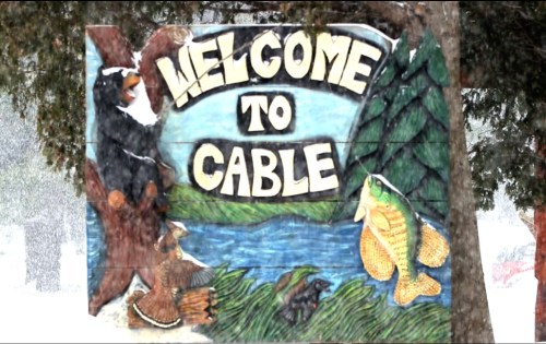 A colorful wooden sign reading "Welcome to Cable," featuring a bear, fish, and natural scenery. Snow is falling.