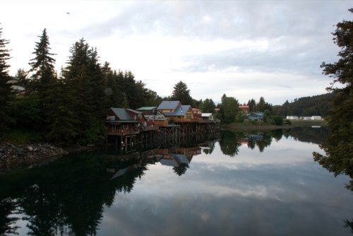 A serene waterfront scene with colorful houses on stilts reflecting in calm water, surrounded by trees and cloudy skies.