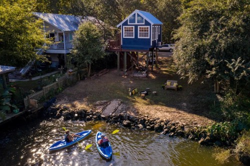 Aerial view of a blue house on stilts by a river, with two kayakers paddling nearby. Lush greenery surrounds the scene.