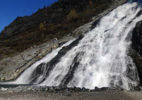 A cascading waterfall flows down rocky terrain, surrounded by greenery and a clear blue sky.