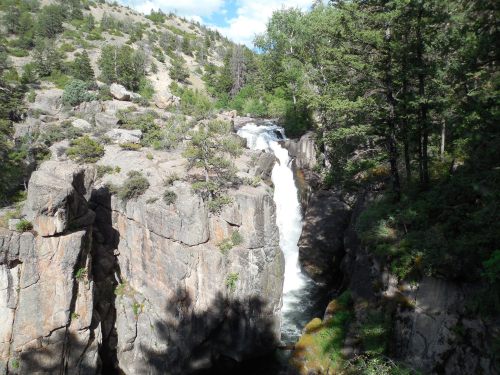 A waterfall cascades between rocky cliffs, surrounded by lush greenery and trees under a bright blue sky.