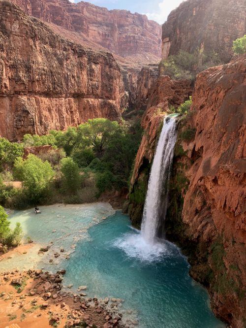 A stunning waterfall cascades into a turquoise pool, surrounded by red rock cliffs and lush greenery.