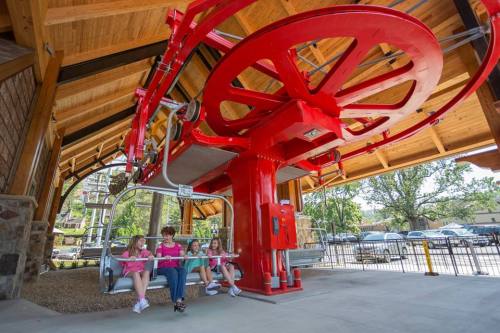A family sits on a ski lift bench under a wooden structure, with a large red wheel overhead and trees in the background.