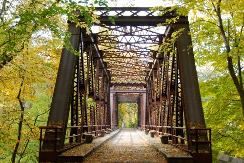 A rusted metal bridge surrounded by vibrant autumn foliage and trees, leading into a serene natural setting.