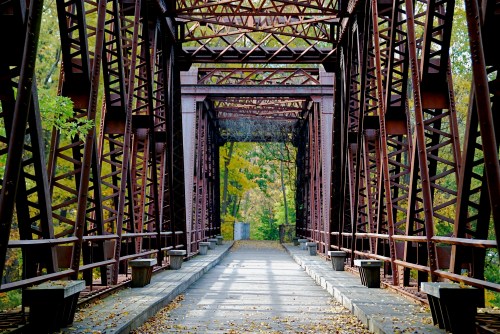A rusted metal bridge surrounded by autumn foliage, leading into a tree-lined path.
