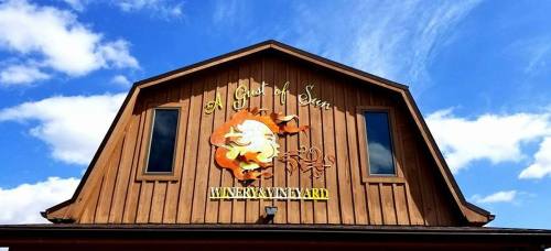 Wooden building with a sign reading "A Gift of Seven Winery Vineyard" against a blue sky with clouds.