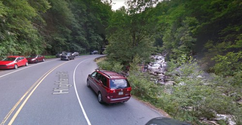 A winding road alongside a river, surrounded by lush green trees and parked cars.