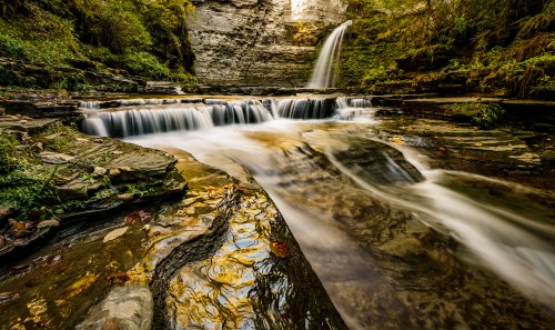 A serene waterfall cascades over rocky ledges into a tranquil pool, surrounded by lush greenery and autumn foliage.