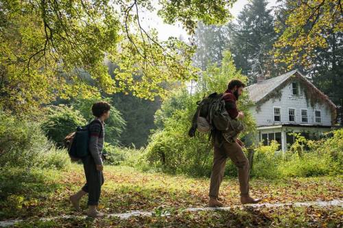 A man and a child walk through a lush, overgrown landscape towards an old, abandoned house.