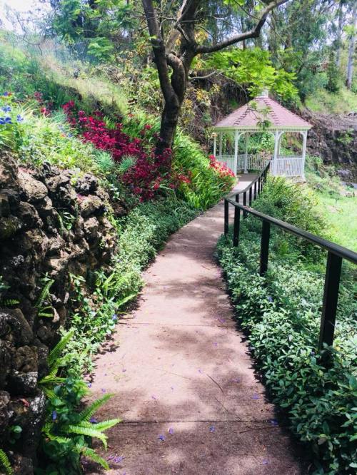 A winding path through lush greenery leads to a white gazebo surrounded by vibrant flowers and trees.