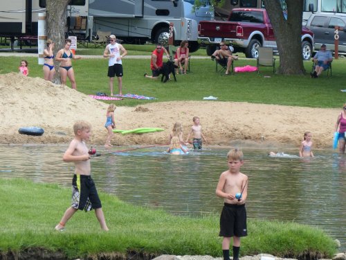 Children play by a sandy beach and water, while adults relax nearby in a park setting.