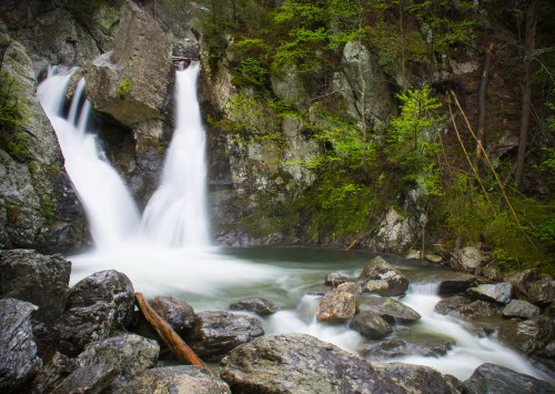 A serene waterfall cascades over rocks into a tranquil pool, surrounded by lush greenery and rugged cliffs.