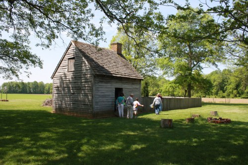 Visitors approach a rustic wooden cabin surrounded by green grass and trees on a sunny day.