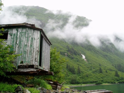 A weathered wooden cabin by a misty lake, surrounded by lush green mountains and low-hanging clouds.