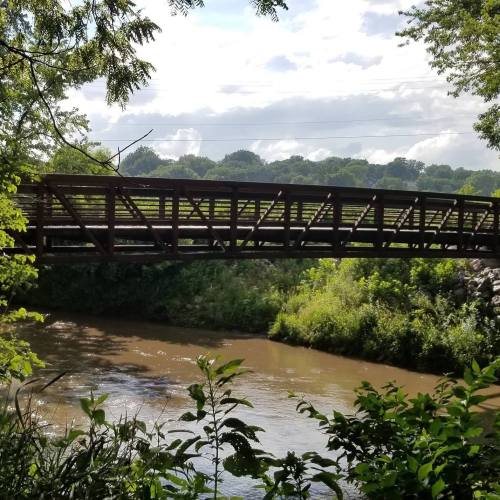 A wooden bridge spans a calm river, surrounded by lush greenery and trees under a partly cloudy sky.