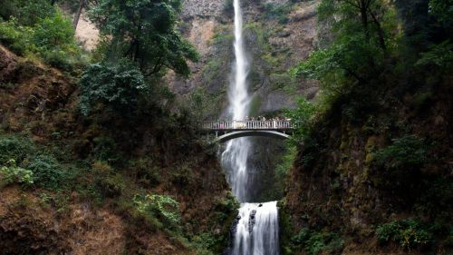 A scenic waterfall cascading down rocky cliffs, with a bridge crossing above, surrounded by lush greenery.
