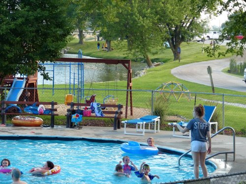 A pool scene with children swimming, a lifeguard on duty, and playground equipment nearby, surrounded by greenery.