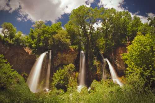 A scenic view of three waterfalls cascading down rocky cliffs, surrounded by lush green trees and a blue sky.