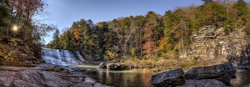 A serene waterfall surrounded by colorful autumn foliage and rocky terrain under a clear blue sky.