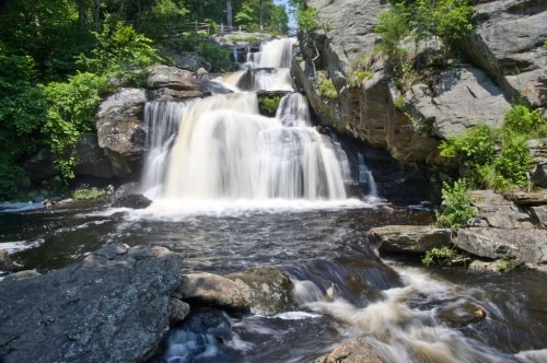 A serene waterfall cascading over rocks, surrounded by lush greenery and a calm river below.