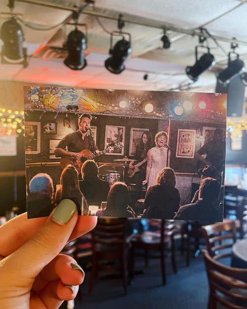 A hand holds a photo of a live music performance in a cozy venue, with musicians and an audience enjoying the show.