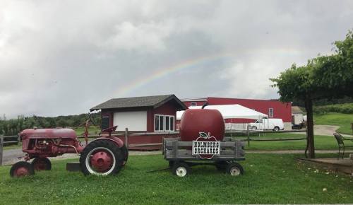 A red tractor and a large apple decoration in front of a building, with a rainbow in the cloudy sky above.