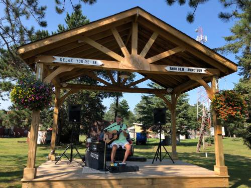 A musician plays guitar on a wooden stage in a park, surrounded by trees and colorful flower baskets.
