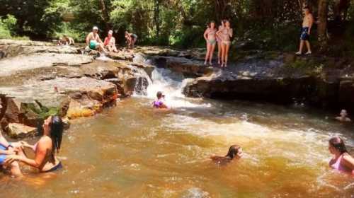 A group of people enjoying a sunny day by a rocky stream, some swimming and others relaxing by the water.