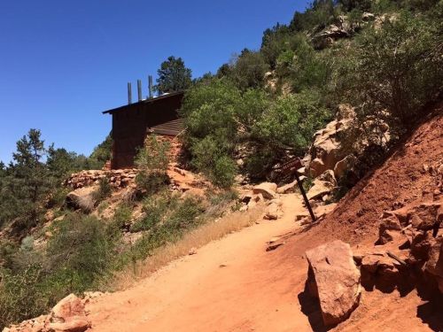 A dirt path leads to a rustic building surrounded by greenery and rocky terrain under a clear blue sky.
