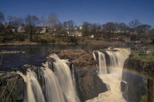 A scenic view of a waterfall with a rainbow, surrounded by trees and buildings under a clear blue sky.